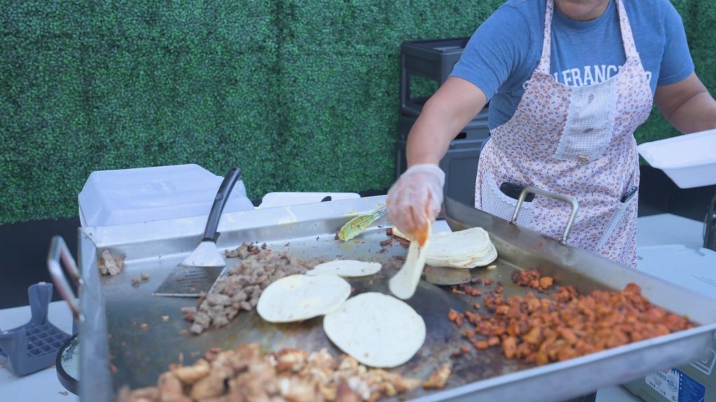 Vendor cooking tacos and warming tortillas on a flat-top griddle at La Fiesta Hispana
