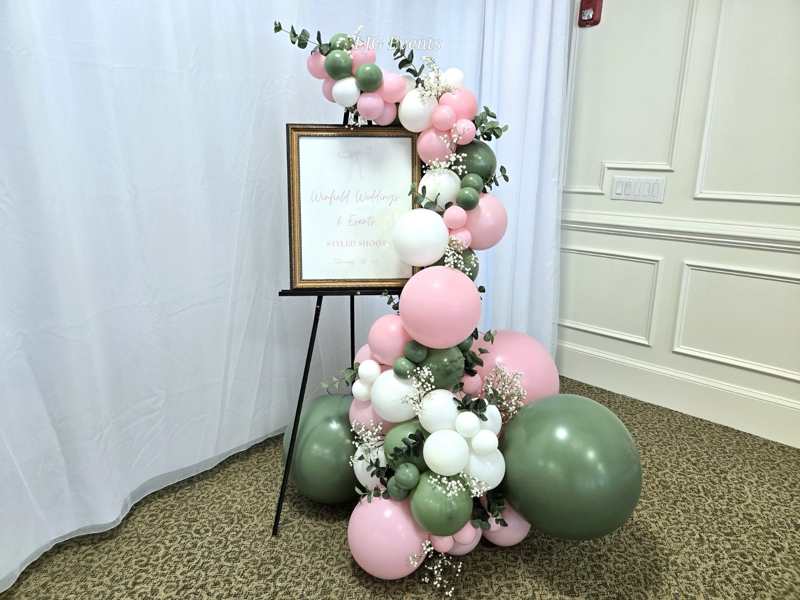 Pink, white and sage balloon garland framing a gold-framed welcome sign on an easel, styled by LJG Events